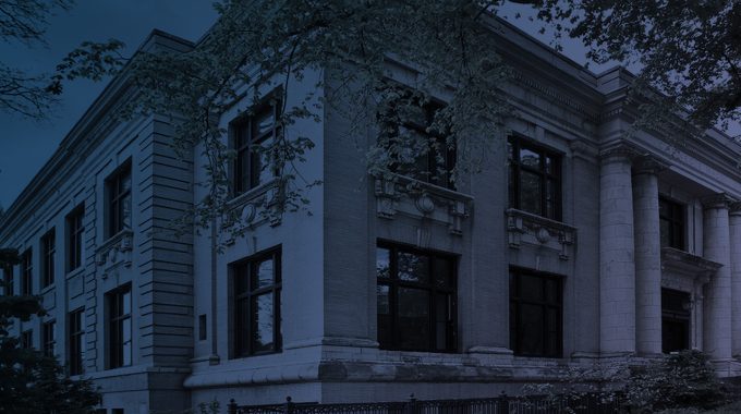 Dark blue and purple tinted image of a two-story stone building with large windows and four pillars framing the entrance