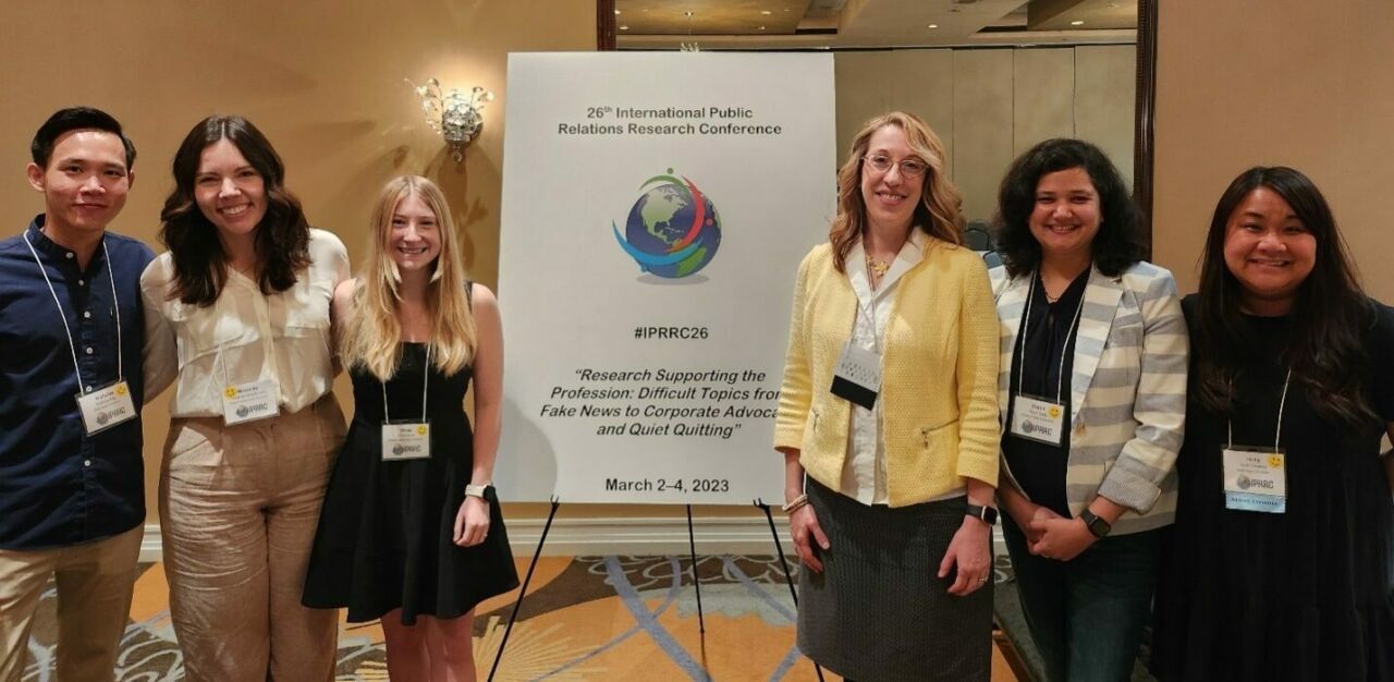 Group of six students and faculty pose in front of a sign that reads 26th International Public Relations Conference